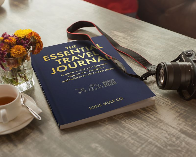 Navy & Gold Essential Travel Journal beside a camera and cup of tea on a table.